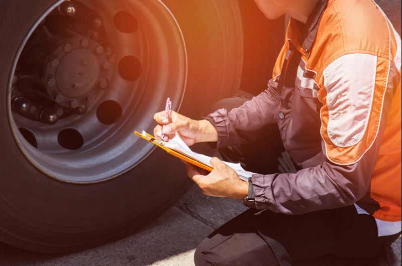 A truck driver holding a clipboard is inspecting a truck's tires.